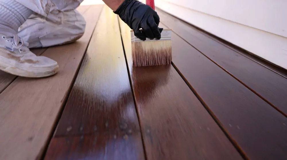 A gloved hand brushing stain onto weathered deck boards